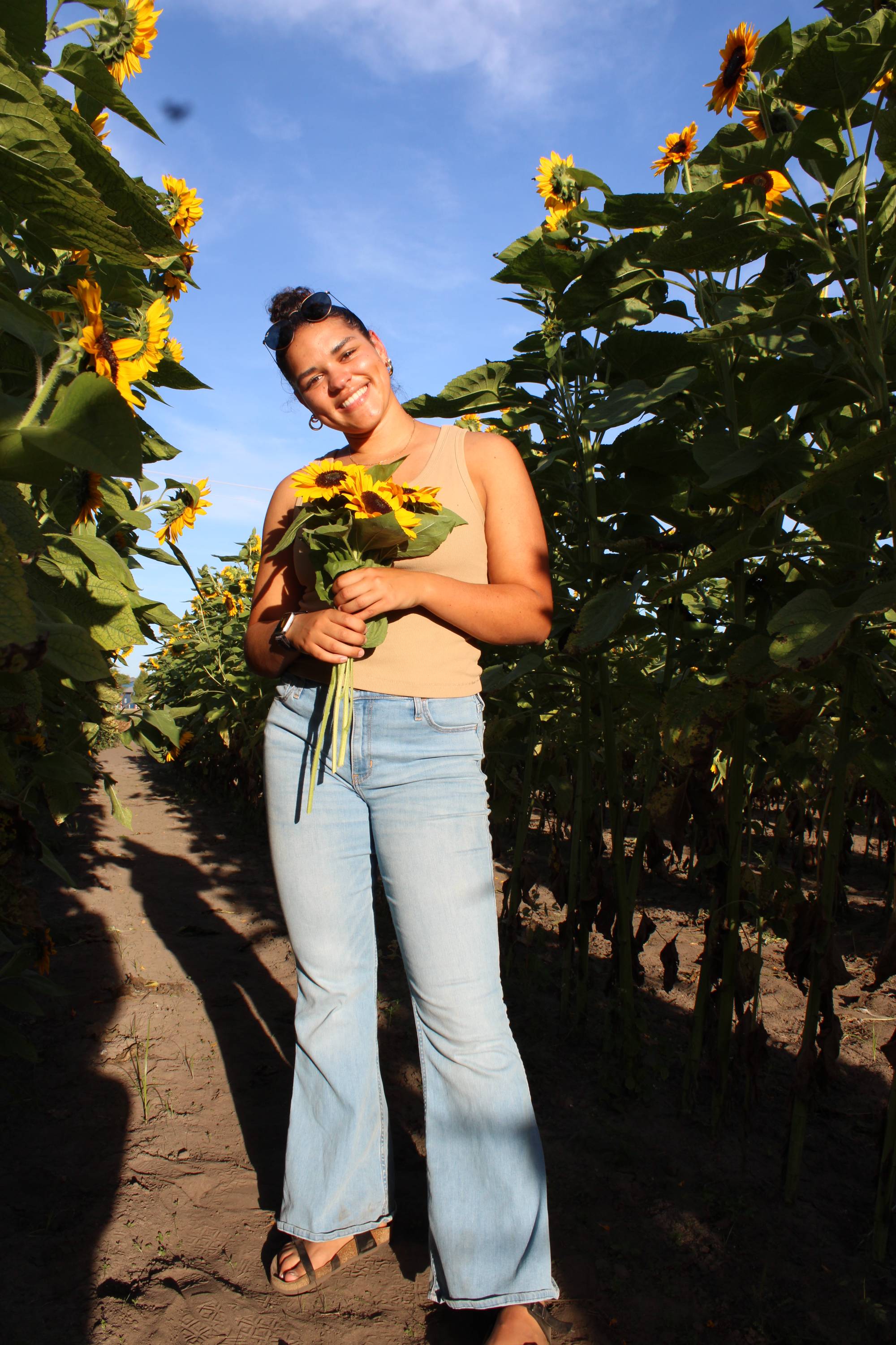 woman with brown hair, a yellow top, and jeans smiling at the camera while holding sunflowers
